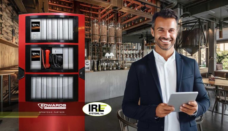 Man in a black blazer holding a tablet in a modern restaurant setting, with a fire alarm control panel labeled "EDWARDS" and "IRL SYSTEMS, Inc" visible, highlighting professional fire alarm solutions in Brooklyn.